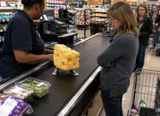 A large cheese block with tiny legs on a grocery conveyor belt.