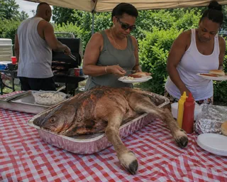 Roasted carcass, fish head, foil pan, checkered cloth, condiments, plates, potato salad, grill, three people, trees.